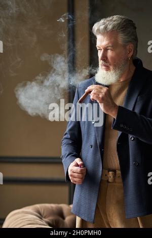 Elégant homme d'âge moyen brutal avec une barbe qui regarde loin tout en fumant un cigare, debout dans un loft intérieur Banque D'Images