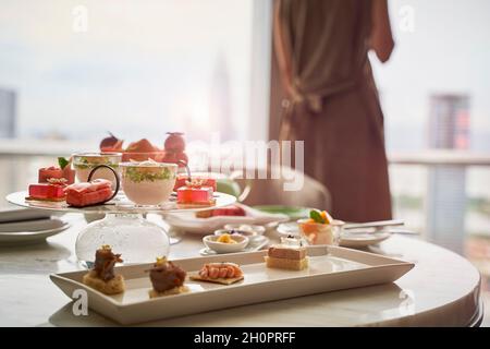 femme asiatique mûre, debout près de la fenêtre, avec vue sur la ville et repas sur table dans une chambre d'hôtel Banque D'Images