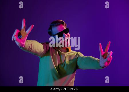 Photo portrait d'un homme souriant portant un verre de soleil montrant le signe de la paix en V souriant isolé sur fond violet foncé Banque D'Images