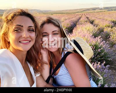 Deux joyeuses femmes amies touristes se tenant debout dans un champ de lavande l'été ensoleillé, souriantes amies se faisant soi-même... Banque D'Images