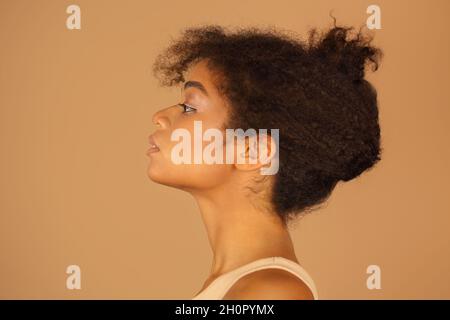 Portrait de profil de beauté de jeune femme afro-américaine confiante avec une peau saine et des cheveux bouclés sur fond brun studio, vue latérale de Banque D'Images