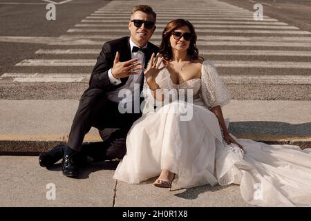 jeunes mariés amoureux, mariés vêcés d'une robe de mariage sont assis sur une traversée piétonne avec toile de fond de paysage urbain et montrent des anneaux de mariage sur f Banque D'Images