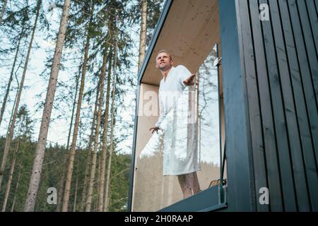Souriant Homme habillé de blanc peignoir debout sur le balcon de la maison de forêt et de profiter de l'air frais avec la nature forêt de pins vues.En dehors de Scandinavian in Banque D'Images