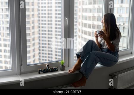 Calme et méditative jeune femme appréciant une tasse de café dans l'appartement loft, tient une tasse de boisson chaude, regarde à travers la fenêtre panoramique et les contempteurs.Matin tranquille Banque D'Images