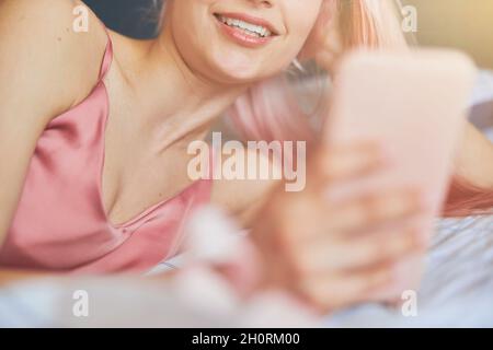 Une jeune femme heureuse en satin rose élégant tient le téléphone sur un lit confortable Banque D'Images