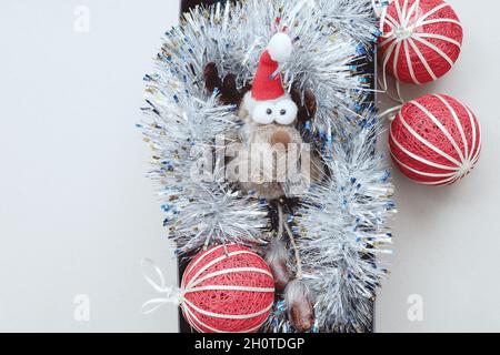 Un petit cerf jouet drôle avec les yeux renflés se trouve dans la guirlande d'argent avec des boules de Noël rouges. Banque D'Images