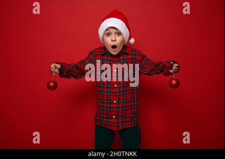 Beau garçon de préadolescence stupéfié, enfant magnifique surpris se tient sur un fond rouge avec des jouets sphériques brillants de Noël dans ses bras, regarde le caméraon Banque D'Images