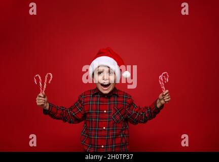 Adorable garçon incroyable, bel enfant pose sur fond rouge coloré avec des cannes de Noël, des sucettes douces rayées dans les mains, des sourires très doux l Banque D'Images