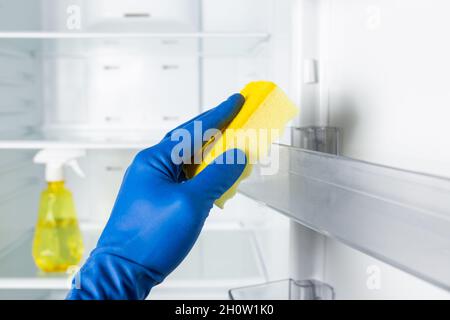 Mains dans des gants de couleur bleue pour nettoyer l'intérieur du réfrigérateur avec un détergent en aérosol et un chiffon. Banque D'Images