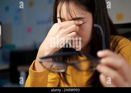Stressée femme épuisée assise au bureau et travaillant des heures supplémentaires, elle est surchargée de travail et de sentir la fatigue oculaire. Banque D'Images