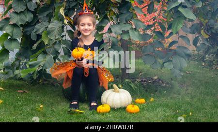 Une fille habillée comme sorcière s'assoit sur une grande citrouille et sourit.Une petite sorcière se cache sous un arbre d'orme de parapluie parmi les citrouilles.Idées de costumes d'Halloween Banque D'Images