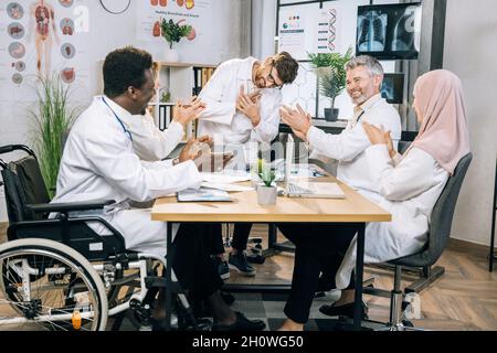 Une équipe de médecins multi-ethniques claquant avec des collègues masculins après un discours pendant la conférence.Des médecins spécialistes heureux avec un en fauteuil roulant assis à la salle de réunion et un collègue. Banque D'Images