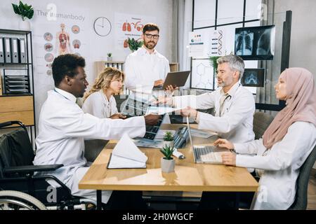 Médecin de sexe masculin debout près d'un mur de verre avec un ordinateur portable dans les mains regardant la caméra.Groupe de médecins multiethniques scientifiques assis à table et de partager des documents et des rayons X.Concept de conférence internationale. Banque D'Images