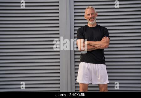 Beau homme d'âge moyen en forme parfaite souriant à l'appareil photo tout en se tenant debout avec les bras croisés à l'extérieur après l'entraînement Banque D'Images