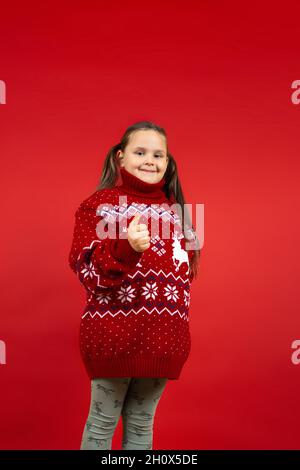 Portrait complet de fille souriante en tricot rouge chandail de Noël avec renne donnant un pouce vers le haut, isolé sur fond rouge Banque D'Images