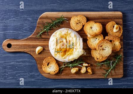 Camembert français cuit au four avec des branches de romarin, des gousses d'ail et des feuilles de Laurier servies avec des croûtons sur une planche de bois sur un bois sombre Banque D'Images