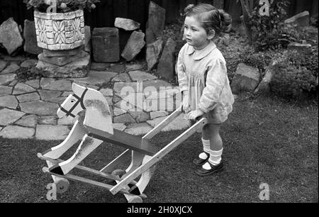 Années 1960, historique, à l'extérieur sur un peu d'herbe par un petit patio, une petite fille debout, tenant la poignée de son jouet porté, une poussée de bois le long de cheval, Angleterre, Royaume-Uni. Banque D'Images