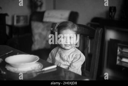 Années 1950, historique, temps de thé et dans une salle à manger, une douce petite fille assise sur une chaise en bois à une table à manger formelle avec un bol vide en face d'elle, Angleterre, Royaume-Uni. Banque D'Images