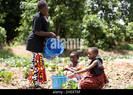 De jeunes femmes africaines noires en difficulté versant de l'eau dans un seau au village bien avec deux petites filles attendant que le récipient soit rempli Banque D'Images