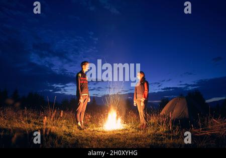 Instantané horizontal de couple de touristes passant du temps ensemble dans le camping.Jeune garçon et fille près d'un feu de camp dans la soirée à l'extérieur dans la nature.Debout regardant le feu dans la nuit d'été Banque D'Images