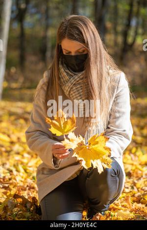 Santé, coronavirus, loisirs, concept de la nature.Brunette femme avec un masque noir sur le visage et un manteau beige ramassant l'érable jaune Banque D'Images