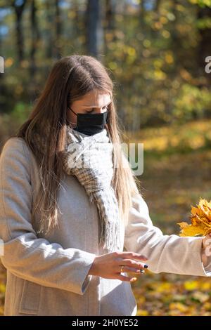 Santé, coronavirus, loisirs, concept de la nature.Brunette femme avec un masque noir sur le visage et un manteau beige ramassant l'érable jaune Banque D'Images