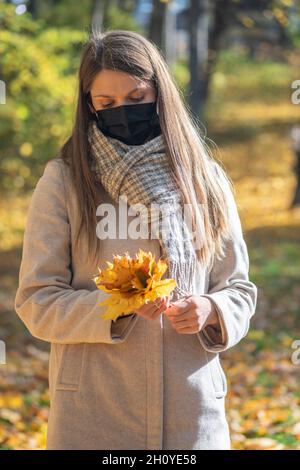 Santé, coronavirus, loisirs, concept de la nature.Brunette femme avec un masque noir sur le visage et un manteau beige ramassant l'érable jaune Banque D'Images