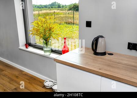 Une bouilloire électrique située sur le comptoir des armoires de cuisine est visible dans la salle à manger avec une grande fenêtre et un vase rouge avec une barre d'or. Banque D'Images