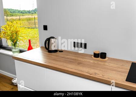 Une bouilloire électrique située sur le comptoir des armoires de cuisine est visible dans la salle à manger avec une grande fenêtre et un vase rouge avec une barre d'or. Banque D'Images