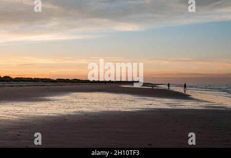 Des figures ombrageuses sur une plage de Norfolk avec des vagues et des reflets aqueux dans la lumière qui s'estompe au coucher du soleil. Banque D'Images