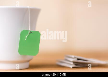 sac à thé avec étiquette verte dans une tasse blanche en céramique sur un plan d'examen, fond jaune chaud, vue rapprochée du modèle de maquette Banque D'Images