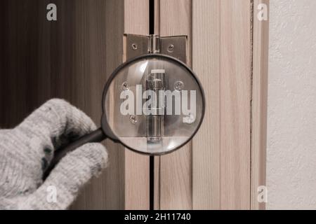 La main d'un ouvrier masculin dans un gant de construction examine à travers une loupe la charnière de porte d'une porte en bois.Le concept d'installation et Banque D'Images