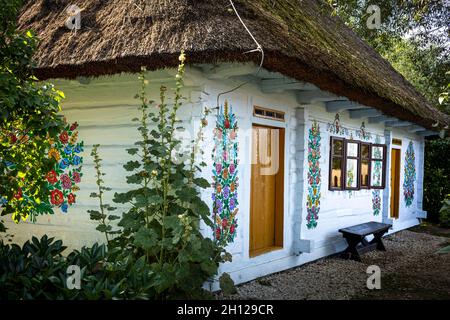 Zalipie, Pologne - 1er août 2021 : chalet traditionnel en bois blanc, avec toit de chaume et murs peints dans un motif floral coloré.Fleurs Hollyhock et un petit banc à l'avant. Banque D'Images