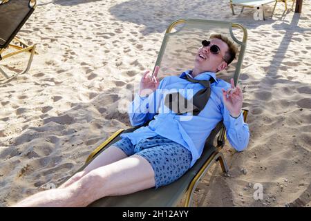 Un jeune homme se trouve sur une chaise longue au milieu de la plage dans une chemise, une cravate et un short de boxer.Affiche les avertisseurs sonores manuels.Aime se reposer Banque D'Images