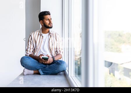 Beau homme indien est assis à la maison sur windowsill, lit des messages texte, envoie des e-mails, utilise son téléphone et une connexion Internet rapide. Banque D'Images