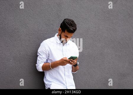 Un homme indien charmant utilise un téléphone portable pour prendre un café contre un mur gris Banque D'Images