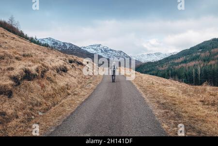 Solitaire routard homme marchant avec sac à dos par Scottish Highlands route de montagne avec des sommets enneigés au début de moody matin.Des personnes actives dans la belle Na Banque D'Images