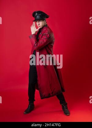 Un jeune homme scandaleux dans un manteau rouge audacieux et dans une casquette en cuir noir, style militaire, dans un style noir vintage, pose épique et fait un geste de main. Banque D'Images