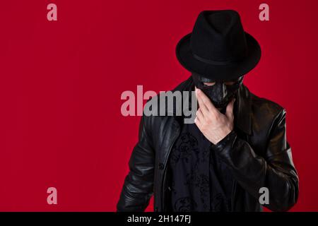 Un mystérieux assassin ninja dans un style noir.Un homme en cuir noir avec un manteau et un chapeau, couvre son visage avec un masque.Photo sur rouge Banque D'Images