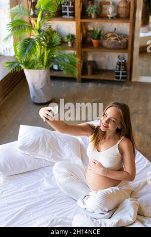Photo d'une jeune femme enceinte s'assoit sur le lit et prend un selfie après se réveiller. Banque D'Images