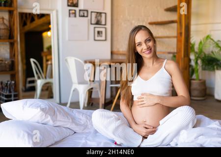 Une jeune femme enceinte est assise sur le lit - elle se réveille, regarde la caméra et sourit.Concept de sommeil sain et de mode de vie actif pendant la prég Banque D'Images