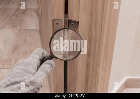 La main d'un ouvrier masculin dans un gant de construction examine à travers une loupe la charnière de porte d'une porte en bois.Le concept d'installation et Banque D'Images