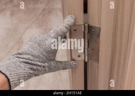 La main d'un homme de travail dans un gant de construction touche et examine une ouverture de porte en bois avec une charnière de porte, gros plan.Installation ou réglage o Banque D'Images