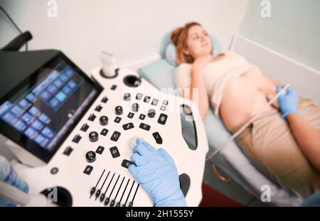 Concentrez-vous sur la main de l'échographiste dans des gants stériles pour l'échographie en clinique.Femme allongé sur un lit de jour alors que le médecin examine l'abdomen du patient avec un échographe sur un arrière-plan flou. Banque D'Images