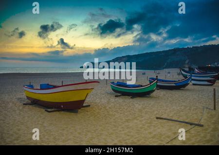 Dessin aquarelle de bateaux en bois colorés sur la plage de Nazaré au crépuscule, bateaux de pêche portugais traditionnels à Nazaré sur la côte, ciel spectaculaire, blanc Banque D'Images