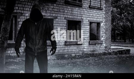 Photo d'horreur effrayante homme de traqueur étranger dans la capuche noire et les vêtements sur le fond abandonné de bâtiment. Banque D'Images