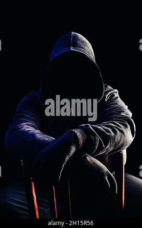 Photo d'horreur effrayante homme de chasse inconnu dans la capuche noire et les vêtements assis sur la chaise sur fond sombre. Banque D'Images