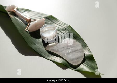 Grattoir de gouache rose, rouleau, pot de crème de feuille verte sur une table blanche. Équipement pour l'auto-massage et les soins de la peau pour le visage et le cou Banque D'Images