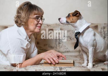 Une femme caucasienne âgée est allongée sur un canapé avec un chien intelligent Jack russell terrier portant des lunettes et une cravate et lisant un livre. Banque D'Images