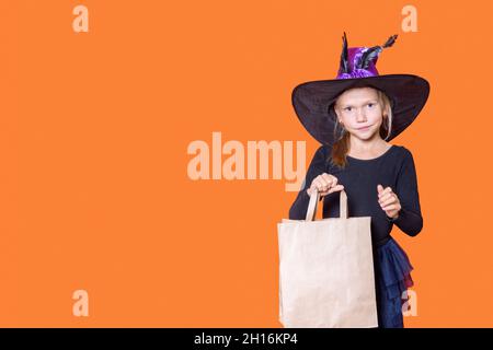 Une belle fille dans une robe noire et un chapeau de sorcière tient un sac de papier d'artisanat, regardant l'appareil photo et souriant, sur un fond orange.Achat de cadeaux c Banque D'Images